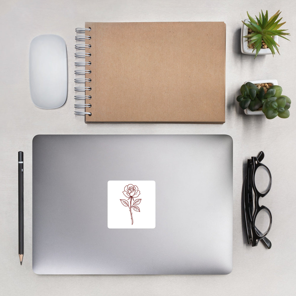 Rose Sticker by By Tee, on a laptop beside a kraft notebook and succulents
