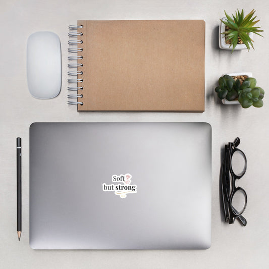 Soft but Strong Sticker by By Tee, on a laptop beside a kraft notebook and succulents