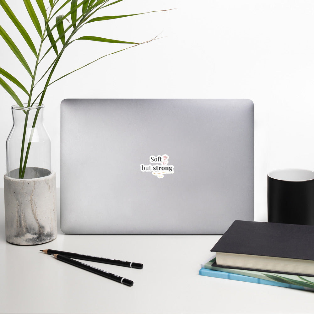 Soft but Strong Sticker by By Tee, on a laptop in a neutral workspace with greenery