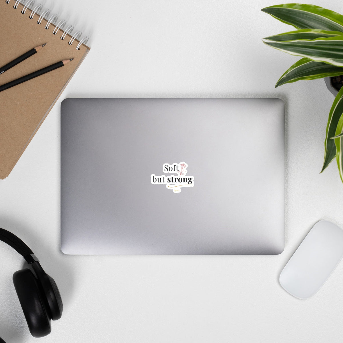 Soft but Strong Sticker by By Tee, on a laptop beside headphones and a plant
